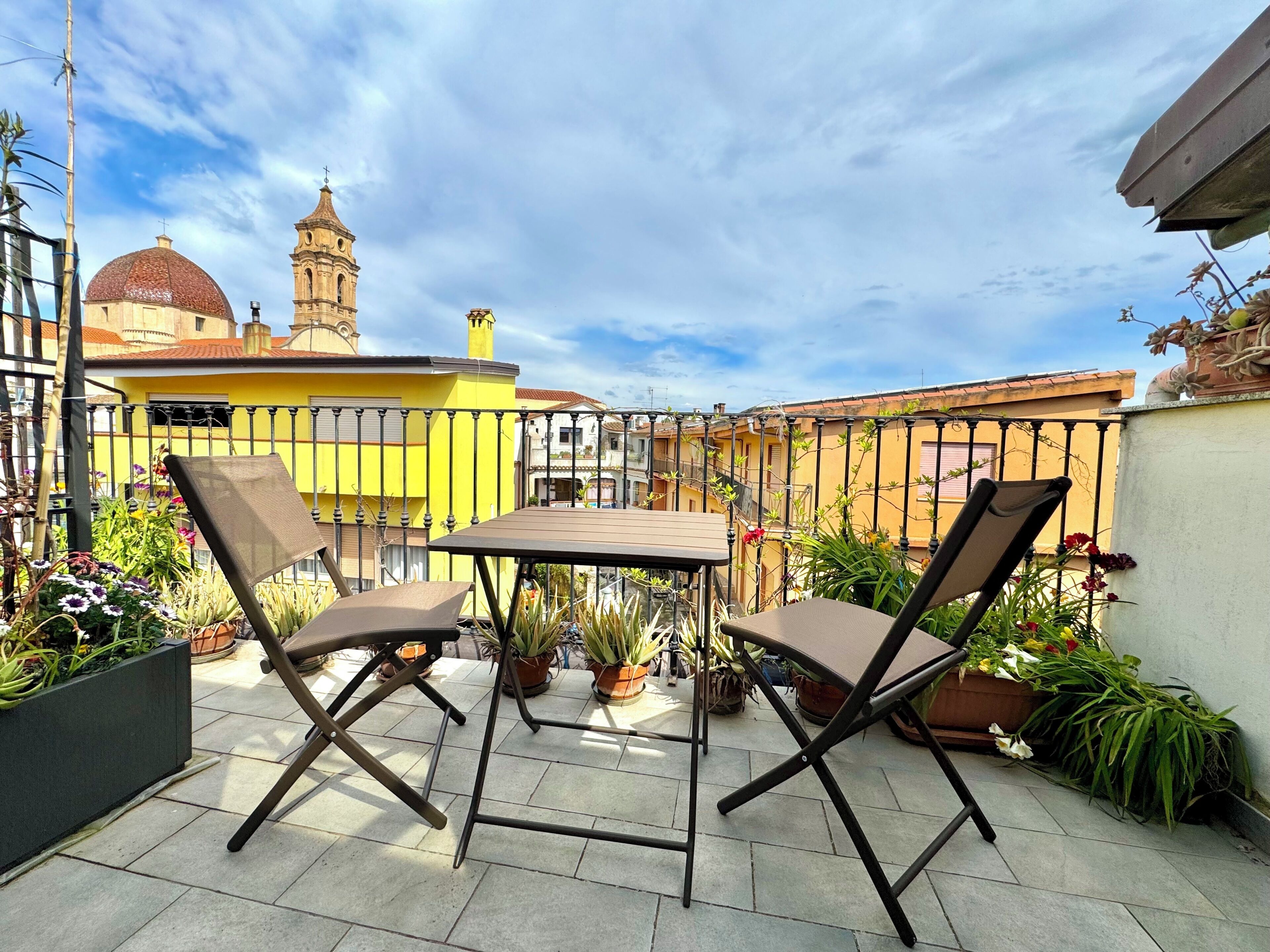 Un patio al aire libre con una mesa y sillas, rodeado de plantas en macetas y un edificio amarillo con cúpula y campanario al fondo.