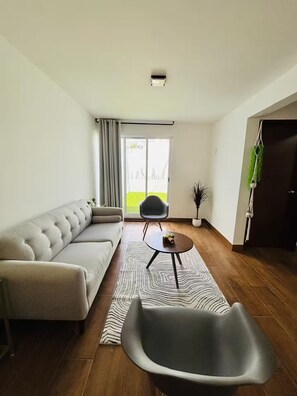 Ein modernes Wohnzimmer mit einer Eckcouch, einem runden Couchtisch und einem einzelnen Sessel. Ein Schiebeglasfenster führt auf einen Balkon.