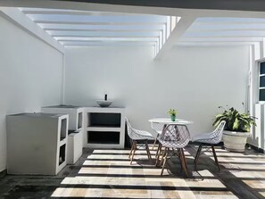 Ein moderner, minimalistischer Außenbereich mit einem weißen Tisch und Stühlen, einer kleinen weißen Kommode und einem Topfpflanzen.