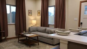 Ein Wohnzimmer mit einer Couch, einem Couchtisch, einer Lampe und einer Küchenzeile mit Arbeitsfläche und Tellern.