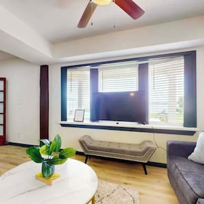 A room with a ceiling fan, a large window with blinds, a grey sofa, a small table with a plant, and a white floor.