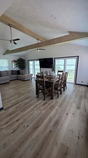 A spacious living area with a dining table set for six, surrounded by wooden chairs. A large flat-screen TV is mounted on the wall, and the room features a high ceiling with exposed wooden beams.