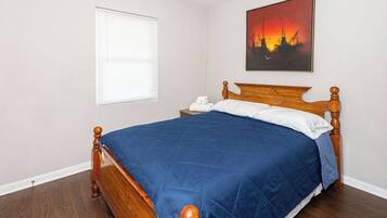 Une chambre à coucher avec un lit en bois, une table de chevet, un oreiller blanc, une couvre-lit bleue, une fenêtre avec des stores et un tableau représentant un coucher de soleil accroché au mur.