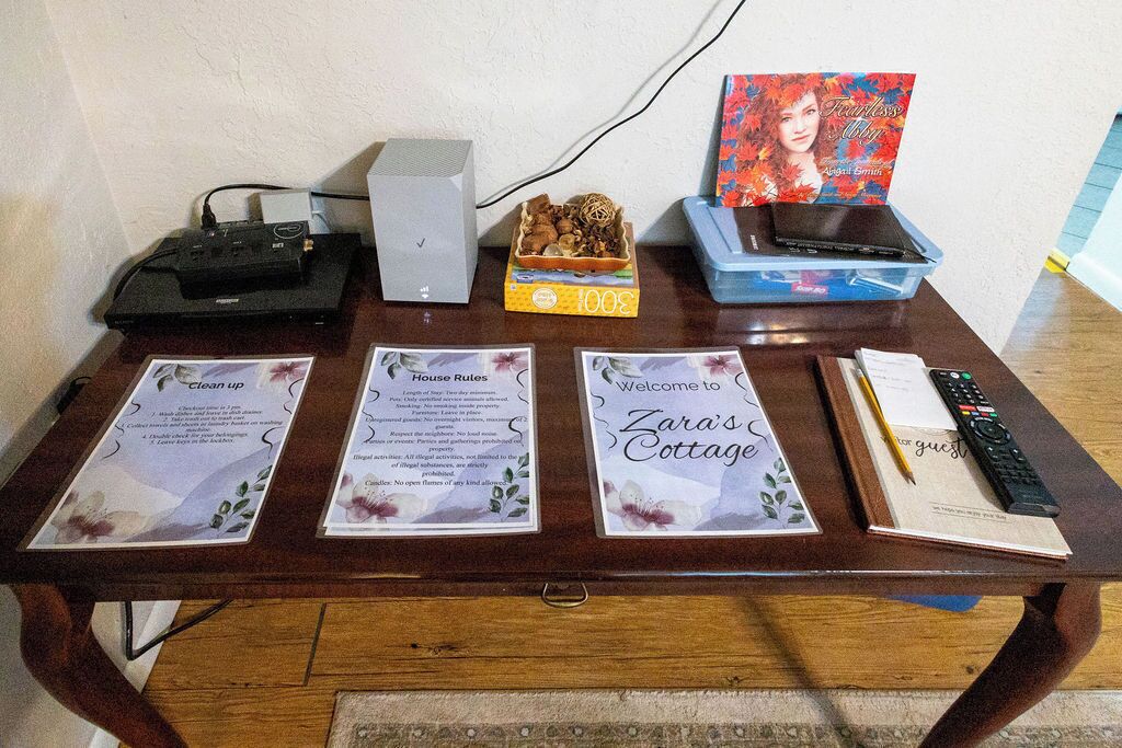 Un tavolo con un telecomando, un libro, una scatola etichettata '300' e tre biglietti informativi intitolati 'Riordina', 'Norme della casa' e 'Benvenuti alla Zara's Cottage'.