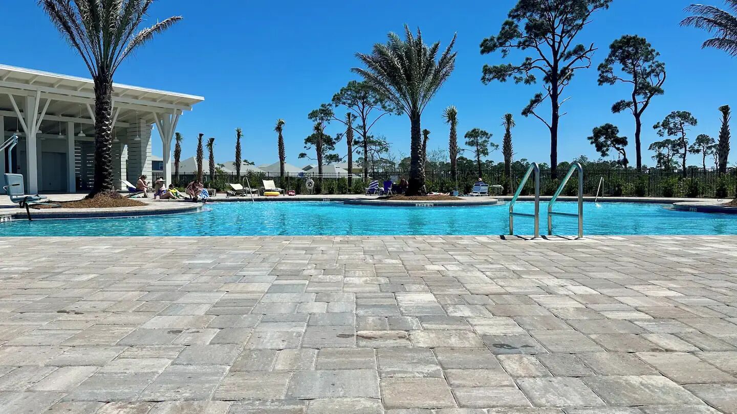 A swimming pool area with a paved deck, lounge chairs, and palm trees.