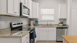A modern kitchen with white cabinetry, a built-in oven, a dishwasher, and a window with blinds. - Golf Cart • EV Charger • Pool • Walk to Beach • 2 Kitchens • Private Suite (Port St. Joe)
