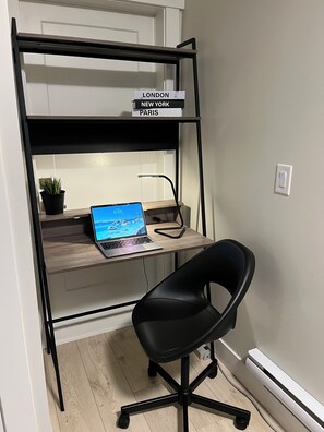 Ein Arbeitsplatz mit Laptop auf einem Schreibtisch, einem schwarzen Bürostuhl und einem Bücherregal mit Büchern mit den Titeln 'LONDON', 'NEW YORK' und 'PARIS'.
