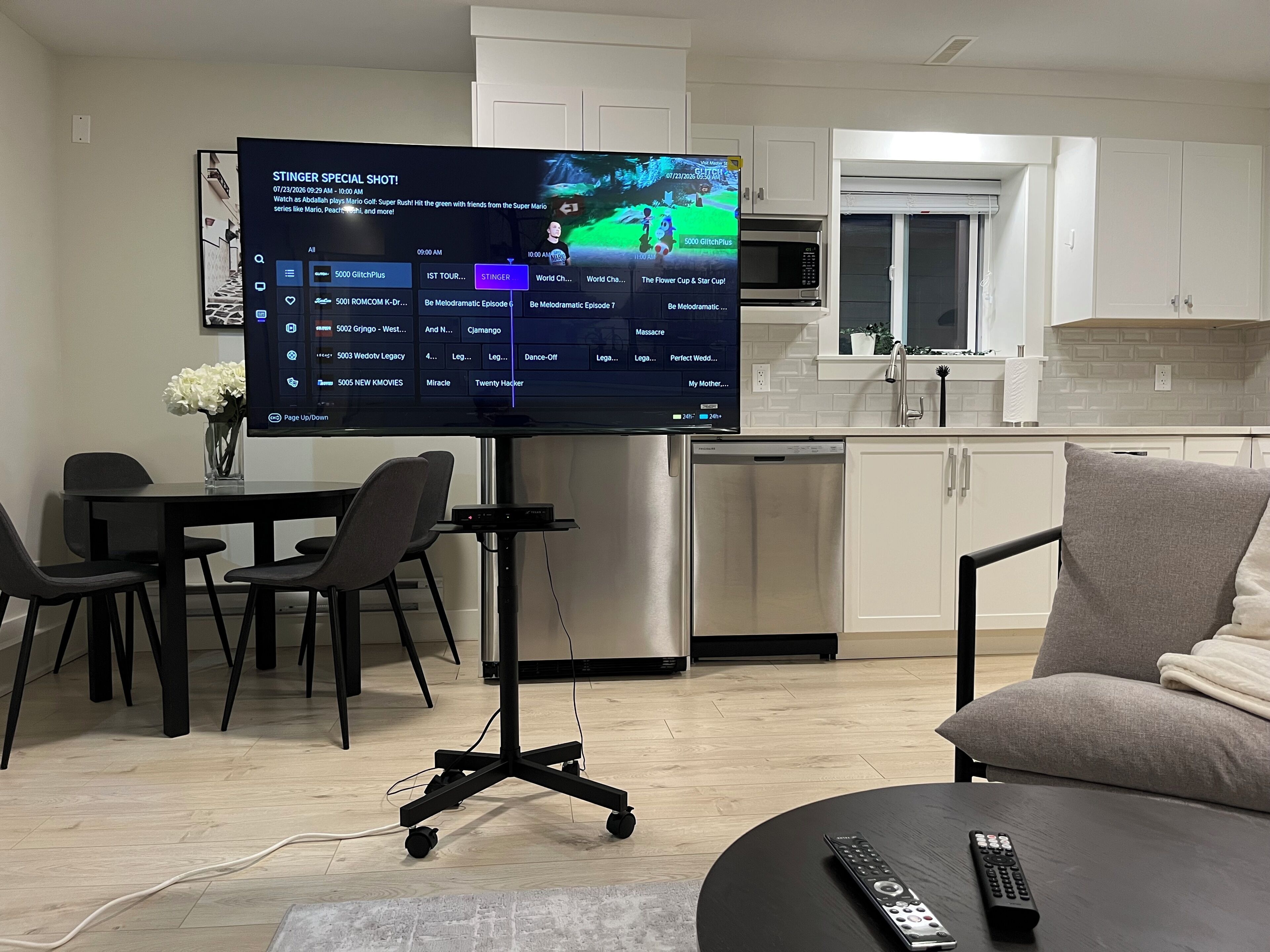 A modern living room with a flat-screen TV mounted on a stand, a dining area with a table and chairs, a kitchen with white cabinets and a microwave, and a grey sofa with a remote control on it.