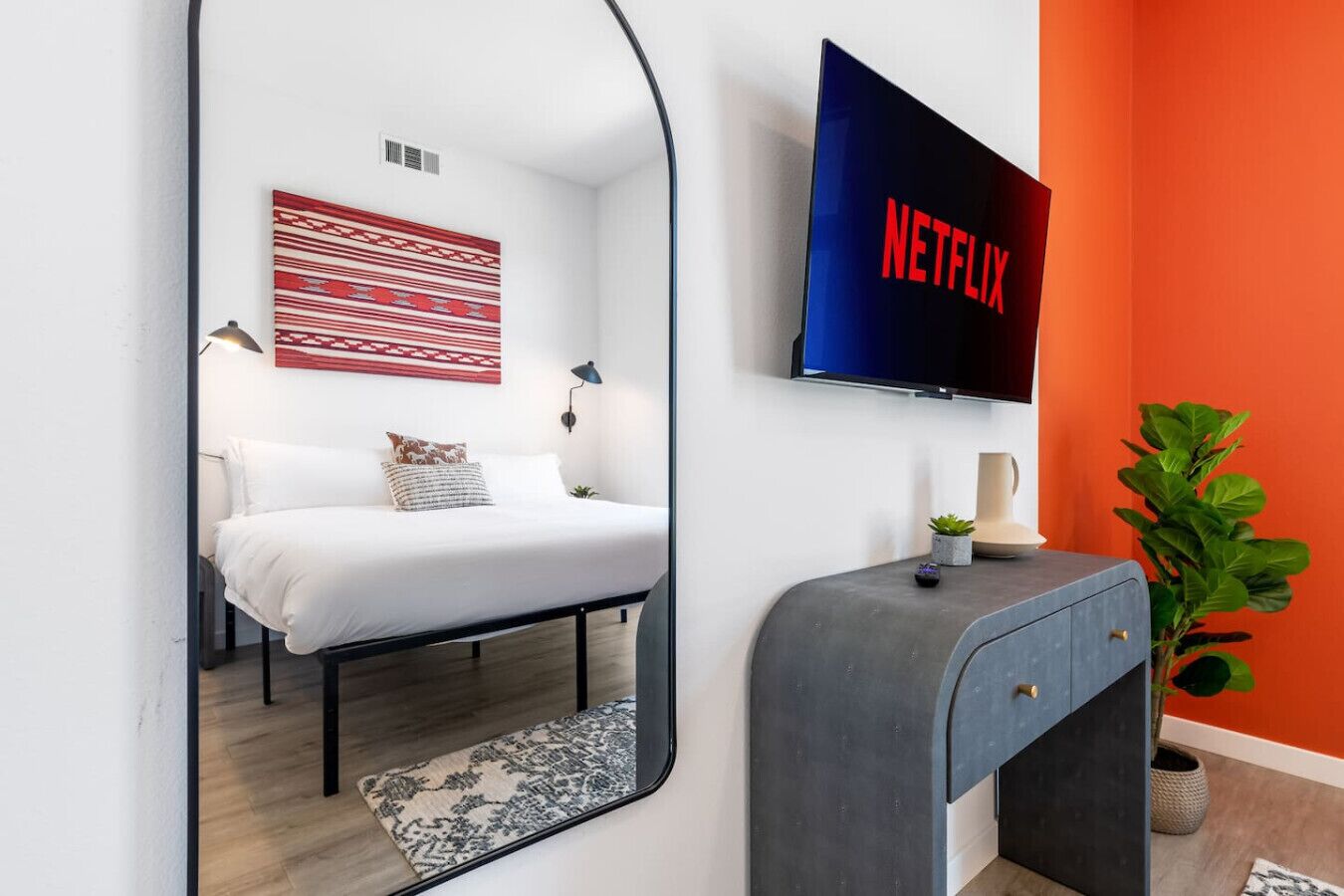 Een moderne slaapkamer met een bed, een aan de muur bevestigde televisie, een nachtkastje en een spiegel die de kamer weerspiegelt.