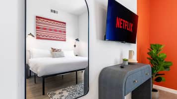 Een moderne slaapkamer met een bed, een aan de muur bevestigde televisie, een nachtkastje en een spiegel die de kamer weerspiegelt.