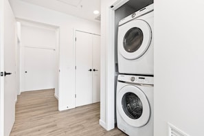 A laundry room with two stacked washing machines and a door leading to another room.