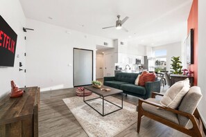 Una sala moderna con sofá, mesa de centro y una silla. Se observa una cocina al fondo con armarios blancos y una zona de comedor.