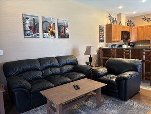 A living room with a black leather sofa, a wooden coffee table, and a kitchen area with wooden cabinets and a refrigerator.