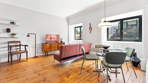 A modern living room with a wooden floor, a red sofa, a dining table with chairs, a TV on a wooden cabinet, and a large window with a view of buildings. - GuestReady - Porto Charming Retreat (Porto)