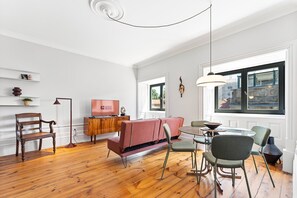 A modern living room with a wooden floor, a red sofa, a dining table with chairs, a TV on a wooden cabinet, and a large window with a view of buildings. - GuestReady - Porto Charming Retreat (Porto)
