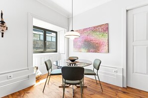 Een eetruimte met een ronde glazen tafel, twee stoelen, een hanglamp en een kleurrijk abstract schilderij aan de muur.