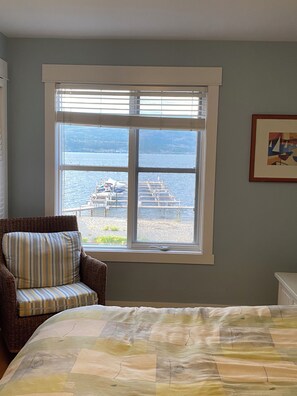 Ein Zimmer mit einem Bett, einem Korbstuhl und einem Fenster mit Blick auf einen Steg und Wasser.