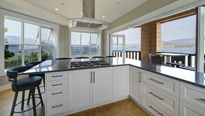 A modern kitchen with a central island, white cabinetry, and large windows offering a view of the sea. - Lakefront Luxury Retreat - Beach & Mountain Views  (Kelowna)
