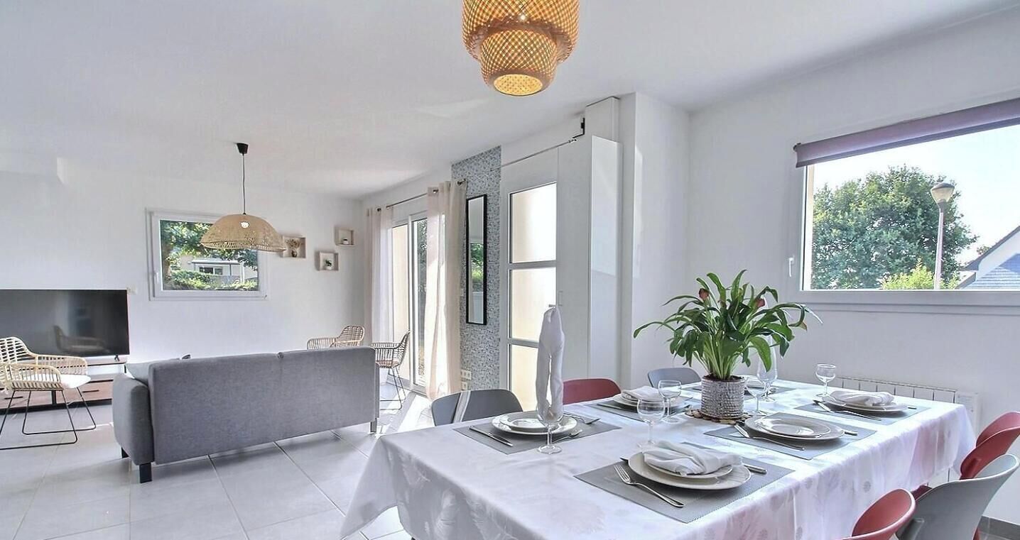Un coin repas avec une table dressée pour quatre personnes, un téléviseur en arrière-plan et une grande fenêtre laissant entrer la lumière naturelle.