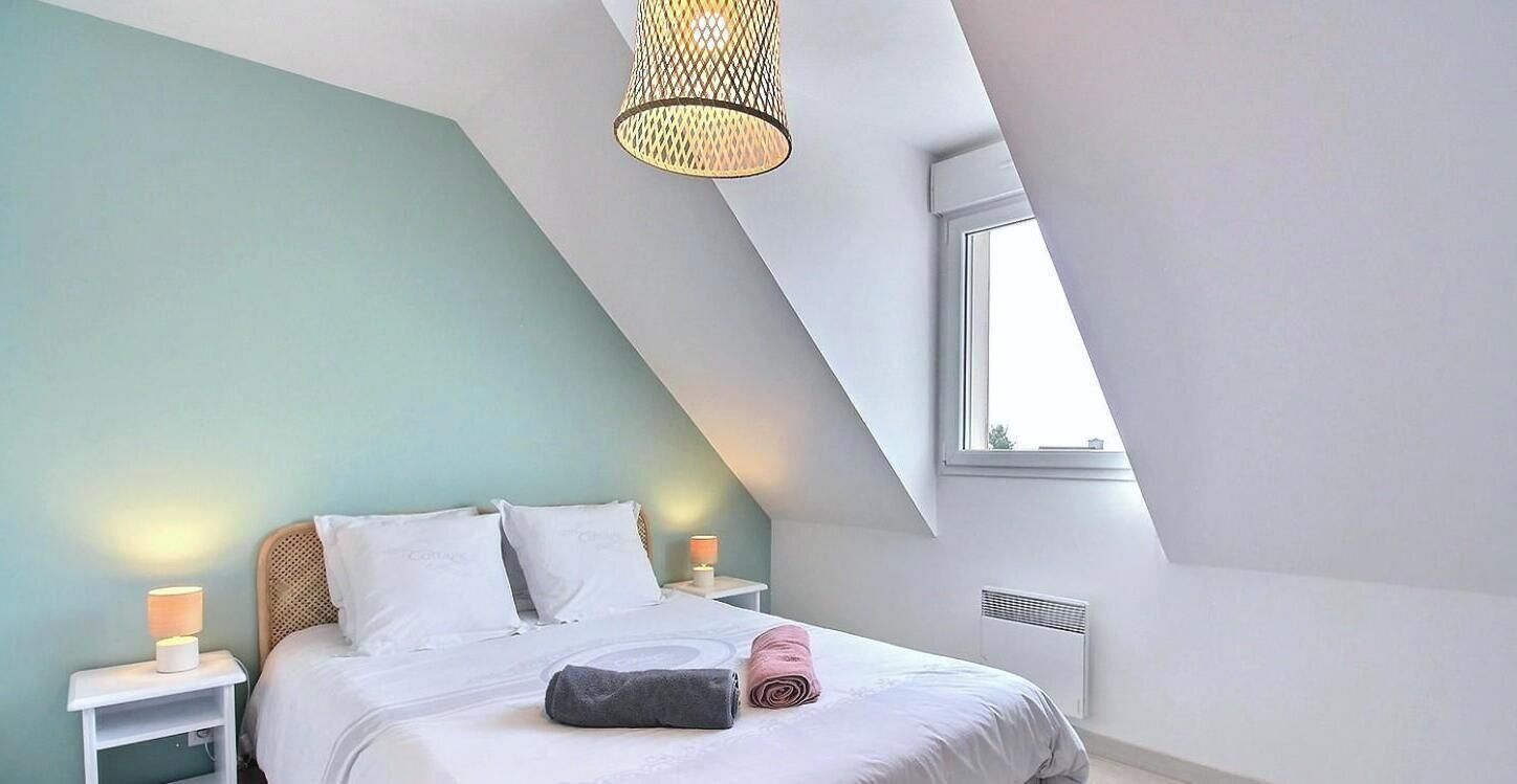 Een slaapkamer met een schuine kap, een bed met witte beddengoed, een bedlampje en een raam.