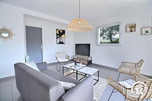 A modern living room with a grey sofa, wicker chairs, a coffee table, a TV, and a large window with a view of trees. - Ty Maezou - LANNION - Vacation Rental (LANNION)