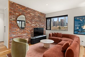A modern living room with a brick wall, a large window with a city view, a flat-screen TV, a sofa, and a coffee table. - Hawke & Hide Designer 3BR Balcony Retreat (West Melbourne)