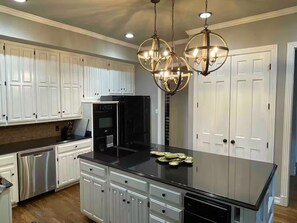A kitchen with white cabinetry, a black countertop, stainless steel appliances, and a chandelier. - Spacious 4 BR Retreat w/Fitness Center & Golf Simulator (Dallas)