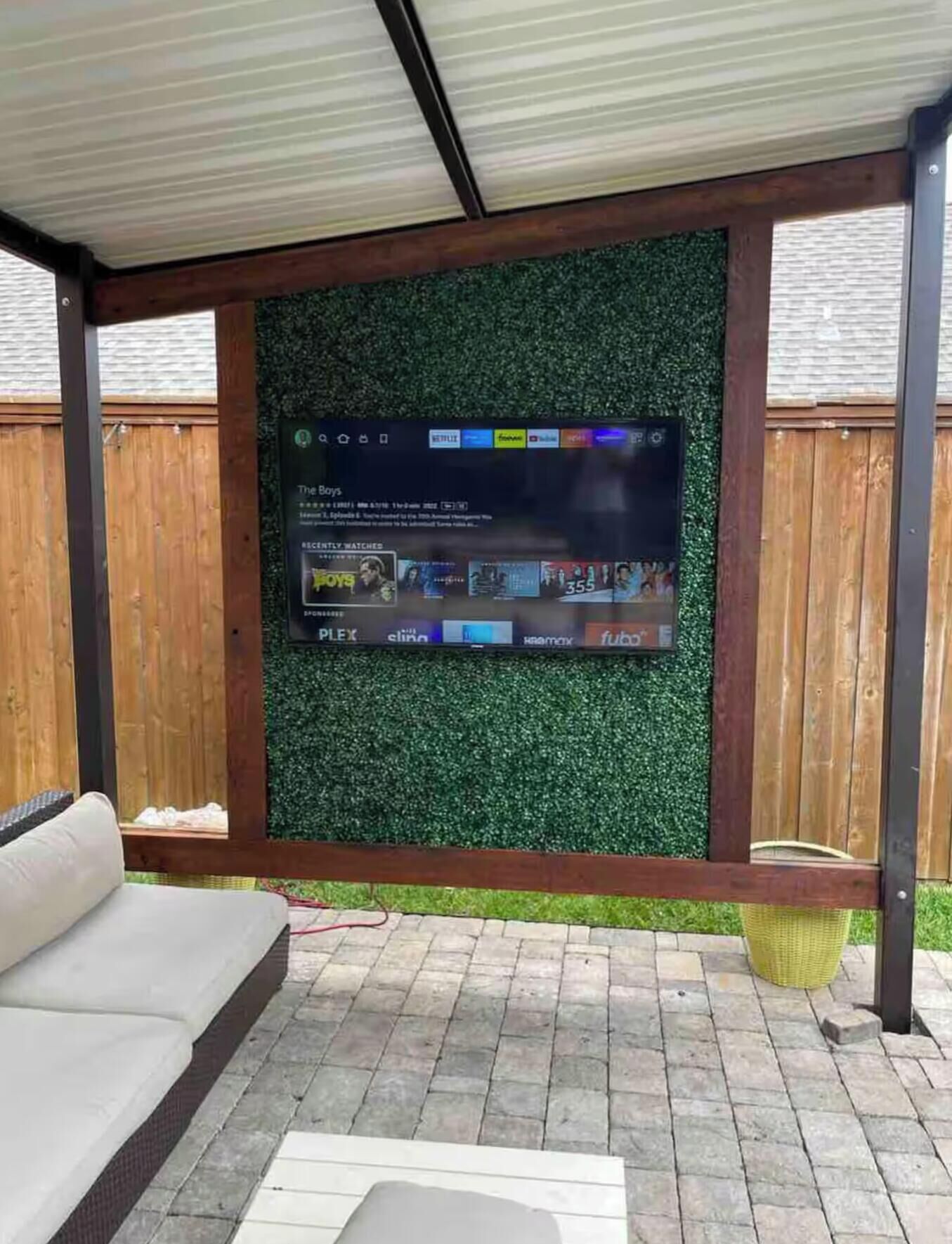 A covered outdoor area with a flat-screen TV mounted on a wall with a green artificial grass backdrop.