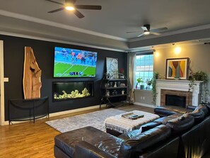 A living room with a leather sofa, a flat-screen TV, a fireplace, and a ceiling fan. - Spacious 4 BR Retreat w/Fitness Center & Golf Simulator (Dallas)