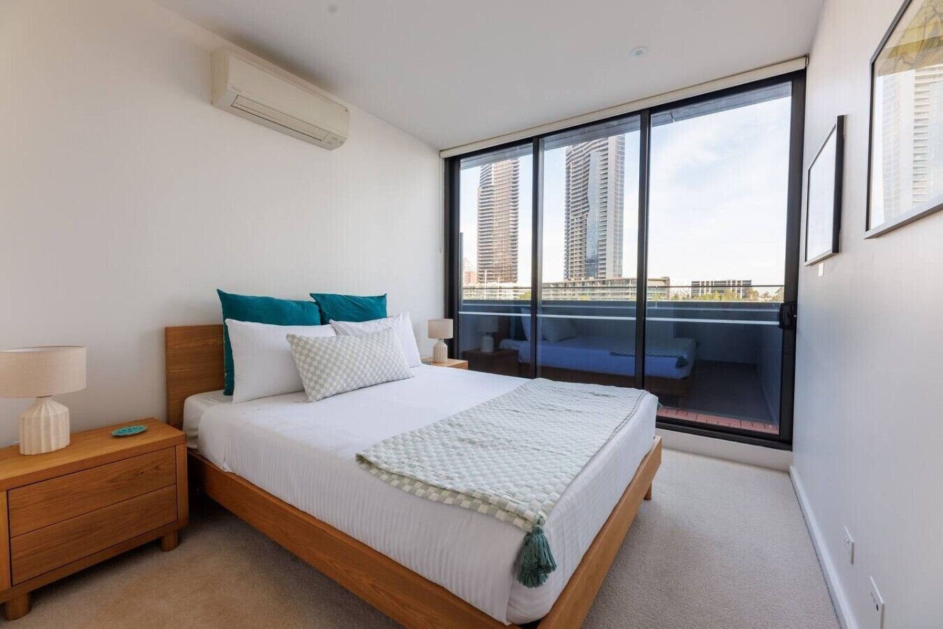 A modern bedroom with a large bed, a nightstand, a lamp, and a view of tall buildings through a large window.