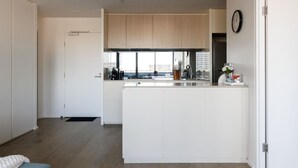 A modern kitchen with a central island, white cabinetry, and a view of the city through large windows. - Marvel & Waterfront Escape - Docklands (Docklands)