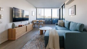 A modern living room with a flat-screen TV, a sofa, a coffee table, and a dining area with chairs and a table. - Marvel & Waterfront Escape - Docklands (Docklands)