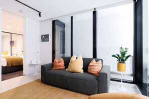A modern living room with a grey sofa, a small white side table, and a potted plant. - Indi | Chic Inner-City Retreat with Parking (Collingwood)