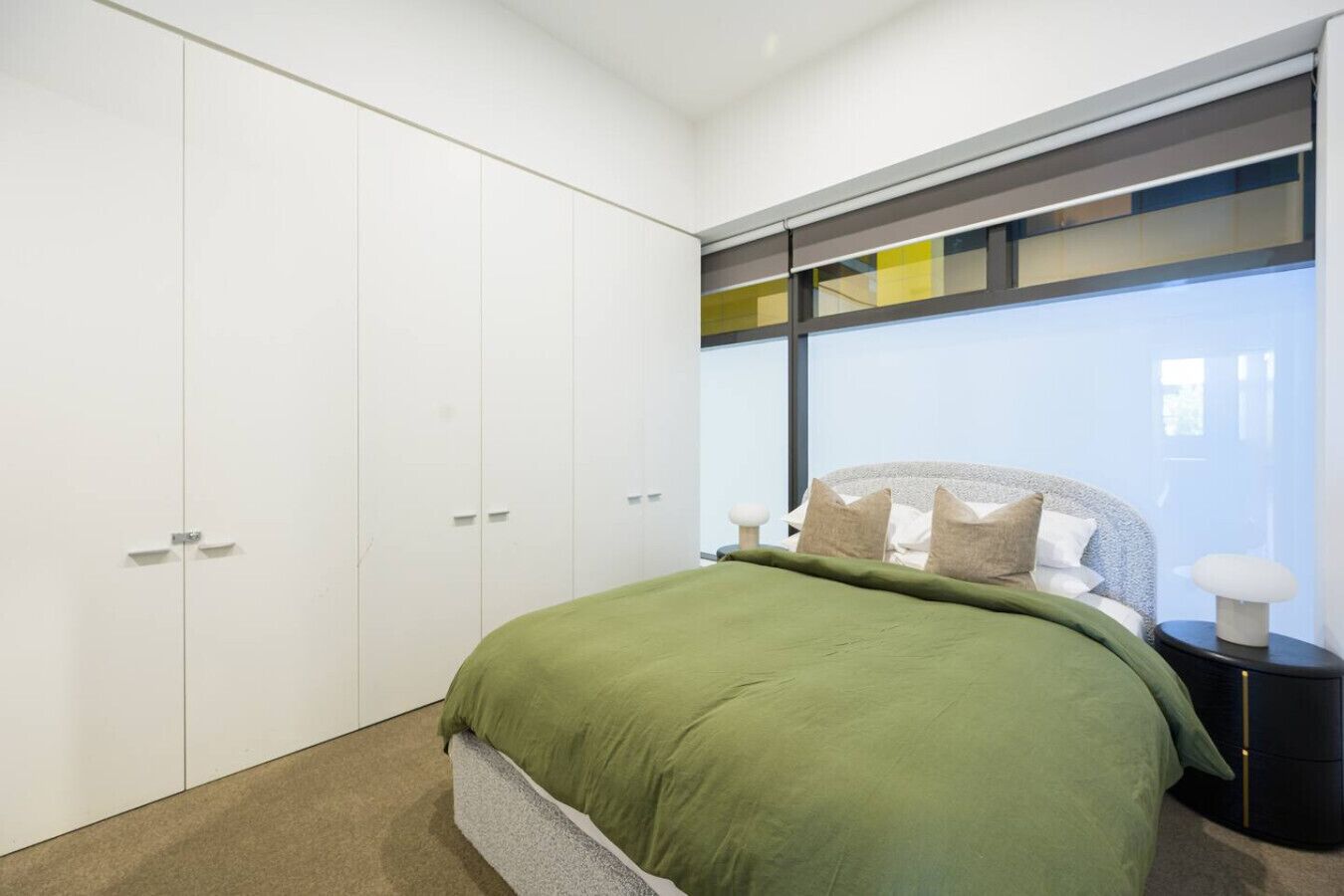 A bedroom with a bed, a nightstand, a lamp, a wardrobe, and a window with blinds.