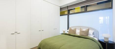 A bedroom with a bed, a nightstand, a lamp, a wardrobe, and a window with blinds.