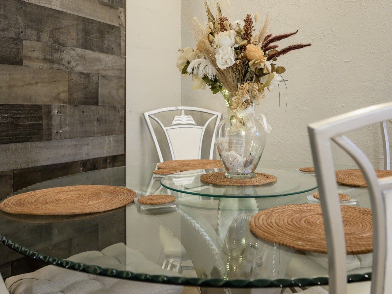 A dining table with a glass top, surrounded by white chairs, and a vase with dried flowers on the table.