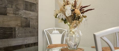 Una mesa de comedor con superficie de vidrio, rodeada de sillas blancas, y un florero con flores secas sobre la mesa.