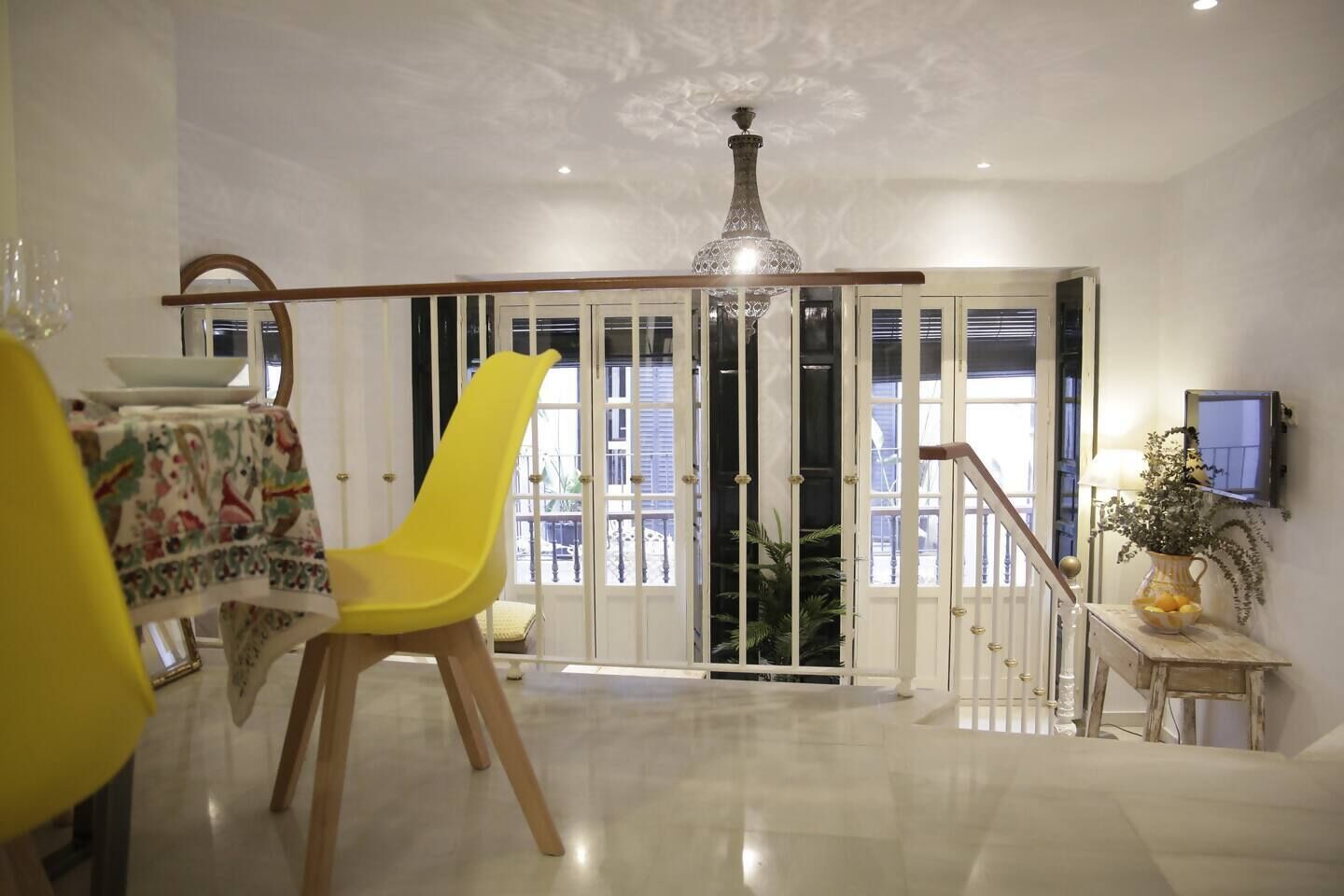Ein geräumiges Zimmer mit Essbereich, ausgestattet mit einem gelben Stuhl und einem Tisch mit gemustertem Tischläufer. Ein großes Fenster bietet Blick ins Freie, und eine Treppe führt zu einer oberen Ebene.