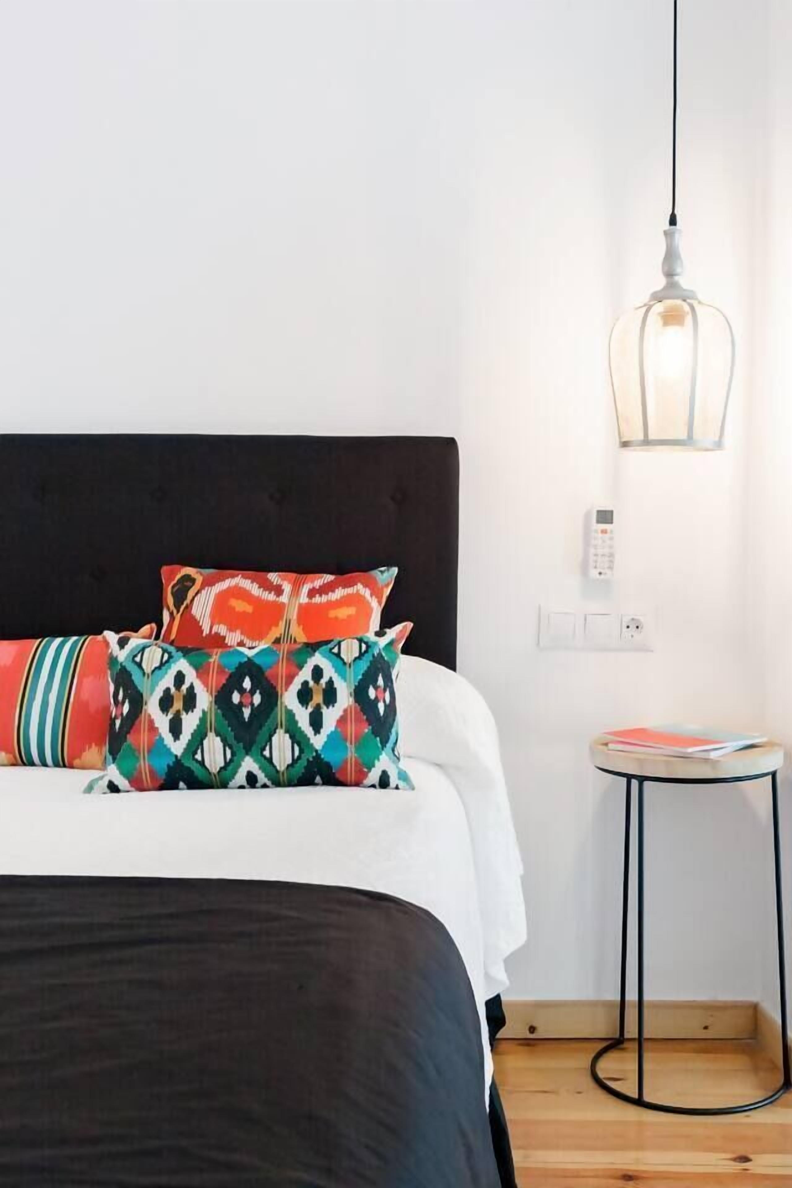 A bed with patterned pillows, a bedside table with books, a wall-mounted light, and a power outlet.