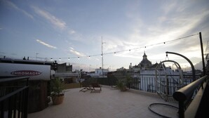 Een terras op het dak met potplanten, zitplaatsen en uitzicht op een historisch gebouw.