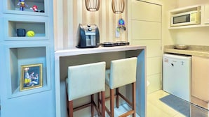 Eine kompakte Küche mit Mikrowelle, Kaffeemaschine und kleinem Kühlschrank. Zwei Barhocker und ein Regal mit Dekorationselementen sind vorhanden.