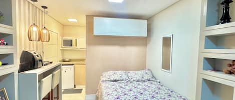 A compact bedroom with a bed, a desk, a microwave, and a small kitchenette.