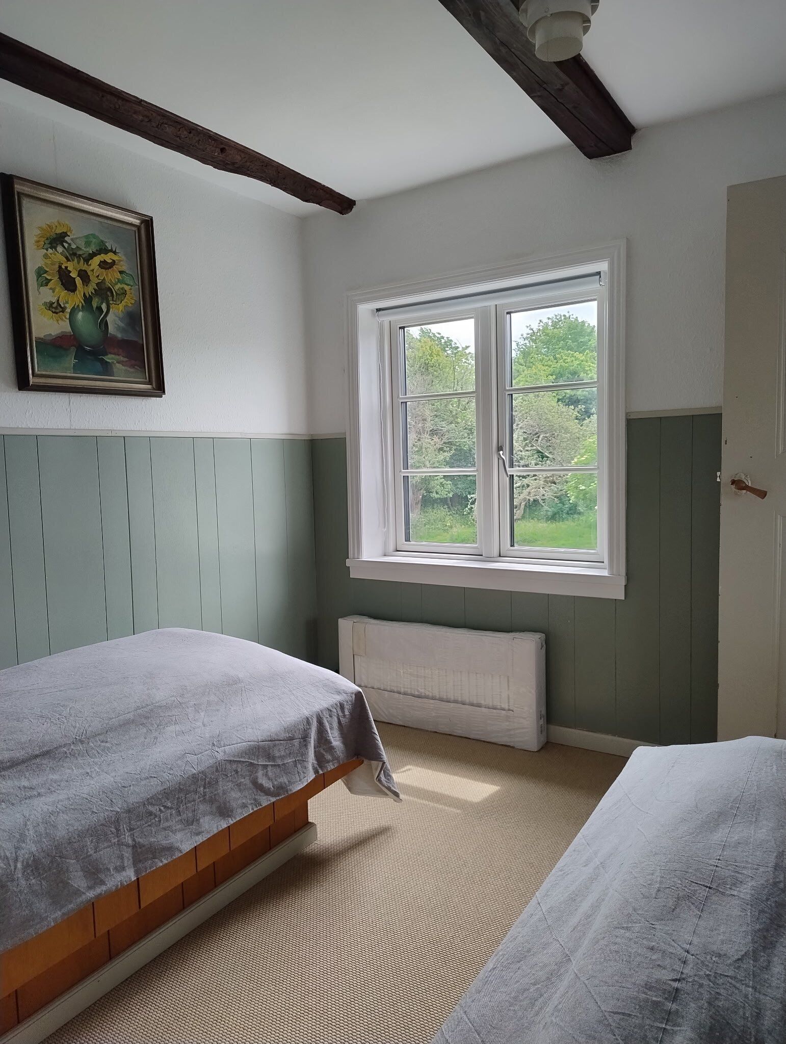 A room with two beds, a radiator, a window with a view of greenery, and a painting on the wall.