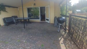 Een overdekt terras met een tafel en stoelen, een barbecue en een schuifpui naar een binnenruimte.