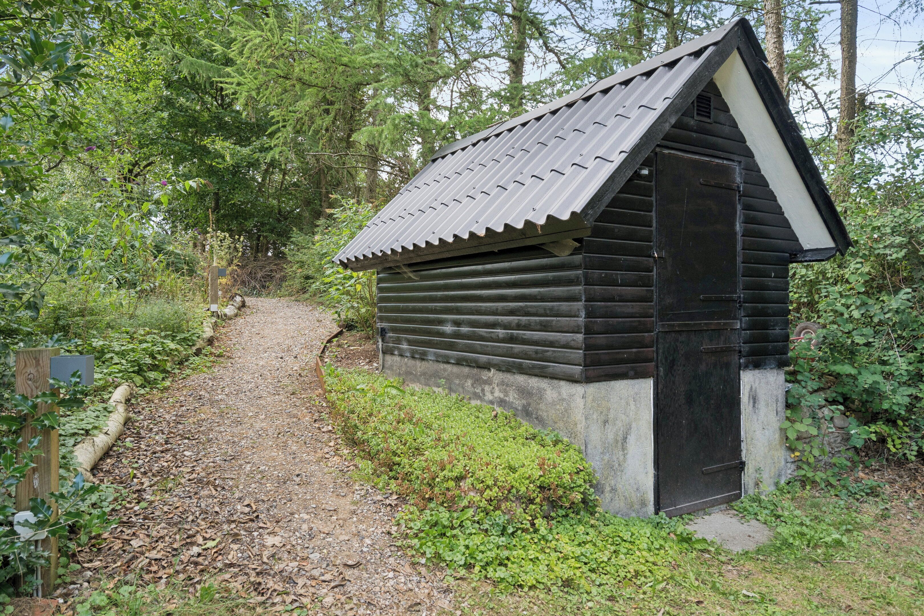 Une petite cabane en bois, dotée d’un toit en pente, située au bout d’un chemin de gravier, entourée d’une végétation luxuriante.