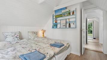 Een slaapkamer met een bed, kussens, een nachtkastje, een wandplank met boeken en decoratieve objecten, en een deur die leidt naar een buitenruimte.