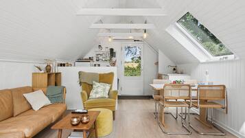A cozy loft-style living space with a high ceiling, a dining area, and a kitchen visible through an open door.