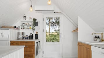 Et køkken med hvide skabe, en sort bordplade, en elkedel og en kaffemaskine. Der er et vindue med udsigt til grøn vegetation udenfor.