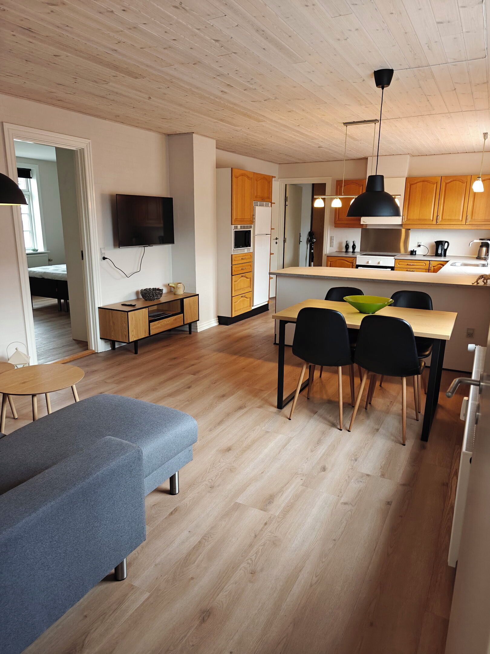A modern kitchen with wooden cabinets, a dining area with a table and chairs, and a living room with a sofa and a TV.