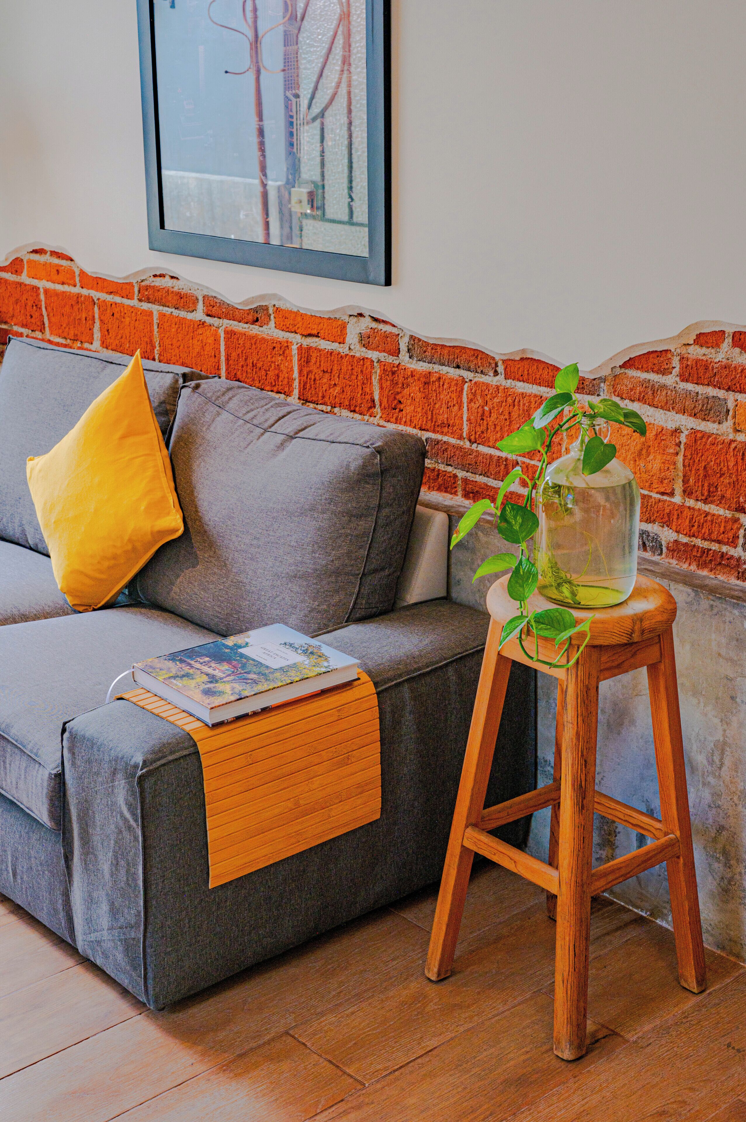 Un salon avec un canapé gris, un coussin jaune, un petit tabouret en bois sur lequel est posée une plante, et un tableau encadré accroché au mur.